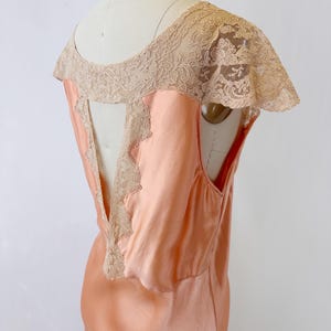 May include: Peach-colored vintage slip dress with a delicate lace overlay on the shoulders and back. The dress features a scoop neckline and a flowing, silky fabric. The lace detailing adds a touch of elegance to this classic garment.