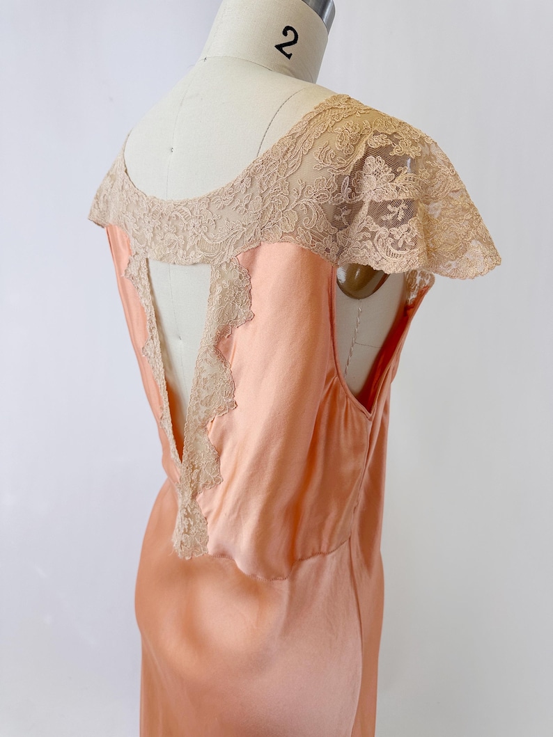 May include: Peach-colored vintage slip dress with a delicate lace overlay on the shoulders and back. The dress features a scoop neckline and a flowing, silky fabric. The lace detailing adds a touch of elegance to this classic garment.