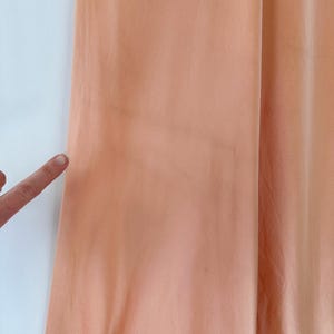 May include: Close-up of a peach-colored fabric, possibly a curtain or garment, with a slight sheen. The fabric is draped, showing soft folds and subtle variations in color. A finger points towards the fabric.