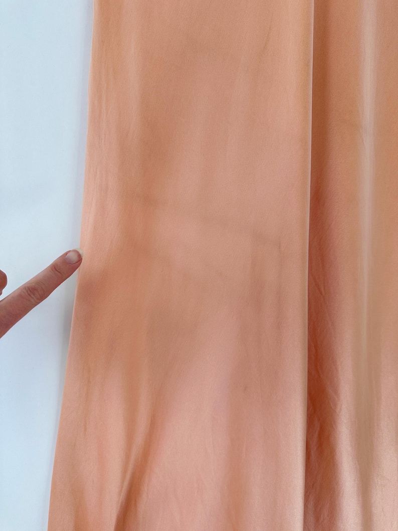 May include: Close-up of a peach-colored fabric, possibly a curtain or garment, with a slight sheen. The fabric is draped, showing soft folds and subtle variations in color. A finger points towards the fabric.