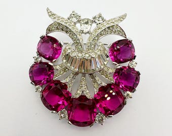 May include: A silver tone brooch with a large, ornate design featuring a cluster of pink rhinestones. The brooch has a fan-shaped design with a central point and a row of rhinestones around the edge.