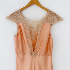 May include: A peach-colored vintage slip dress with a V-neckline and cap sleeves. The dress features intricate beige lace detailing around the neckline and bodice, adding a delicate touch. The dress is hanging on a wooden hanger.