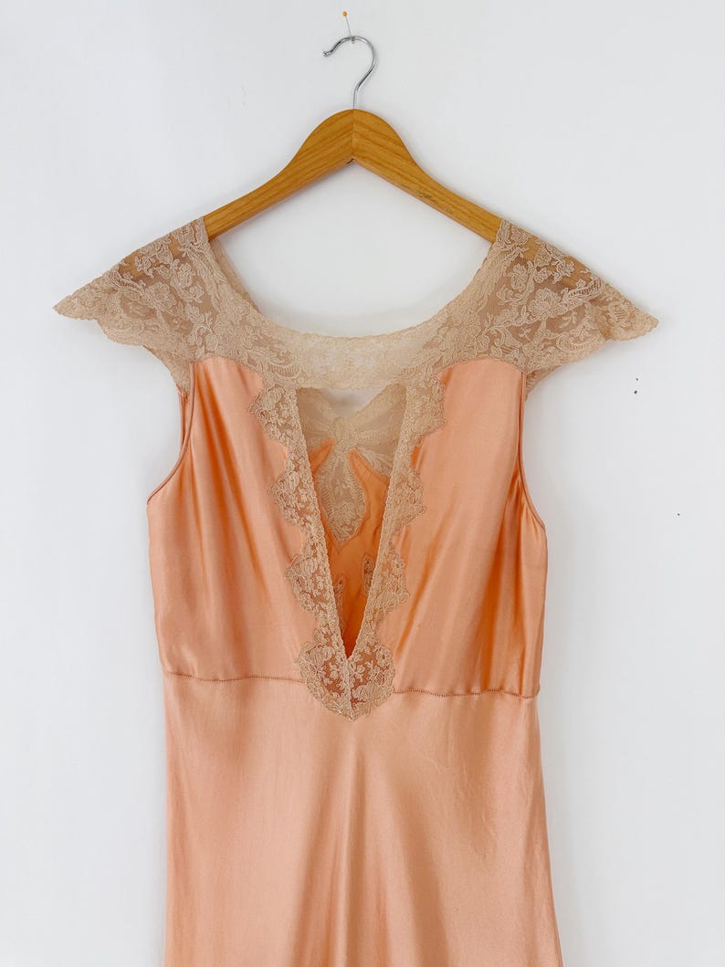 May include: A peach-colored vintage slip dress with a V-neckline and cap sleeves. The dress features intricate beige lace detailing around the neckline and bodice, adding a delicate touch. The dress is hanging on a wooden hanger.