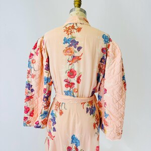 May include: A peach-colored robe with a floral pattern in red, blue, and purple. The robe has quilted sleeves and a matching belt. The floral design is spread across the robe's surface.