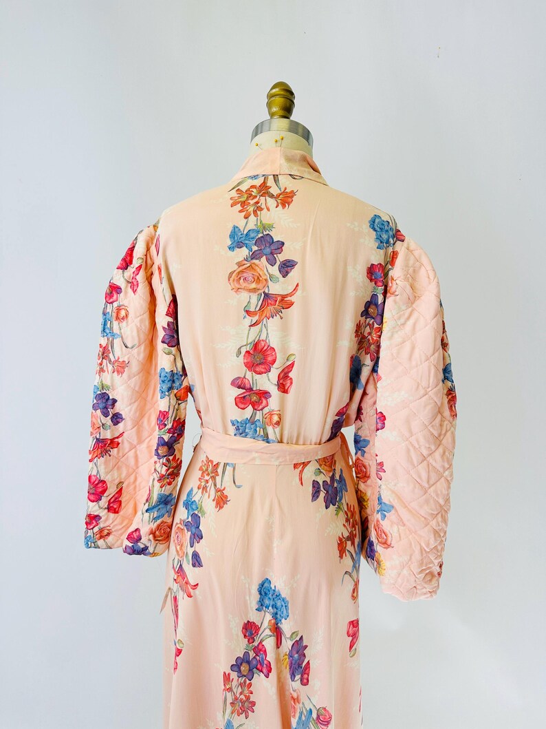 May include: A peach-colored robe with a floral pattern in red, blue, and purple. The robe has quilted sleeves and a matching belt. The floral design is spread across the robe's surface.