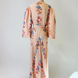 May include: A vintage peach-colored robe with a floral pattern in red, blue, and purple. The robe features quilted sleeves and a matching belt. The long, flowing design is displayed on a mannequin.