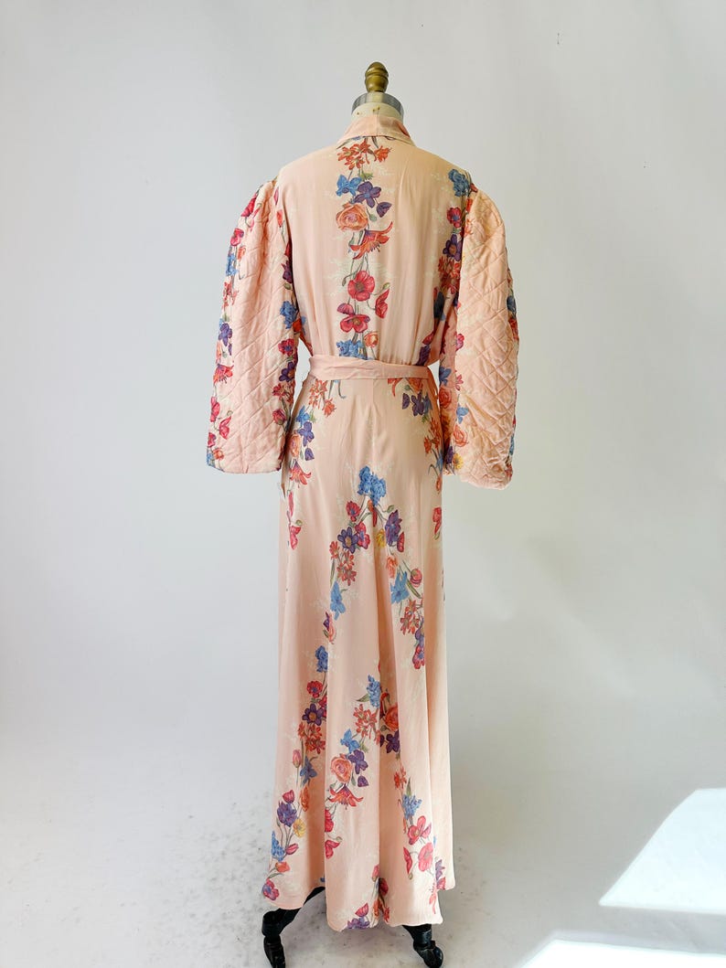 May include: A vintage peach-colored robe with a floral pattern in red, blue, and purple. The robe features quilted sleeves and a matching belt. The long, flowing design is displayed on a mannequin.