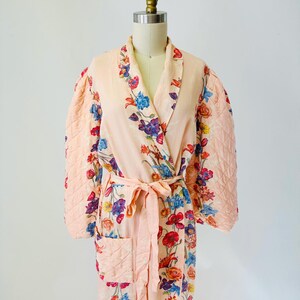 May include: A peach-colored robe with a floral pattern of blue, red, and purple flowers. The robe has quilted sleeves and a matching belt. The robe is displayed on a mannequin.