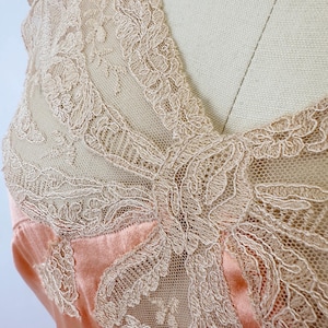 May include: Close-up of a peach-colored satin garment with intricate, cream-colored lace detailing. The lace overlay features floral and geometric patterns, creating a delicate and elegant texture. The garment appears to be a top or dress, showcasing a vintage or romantic aesthetic.