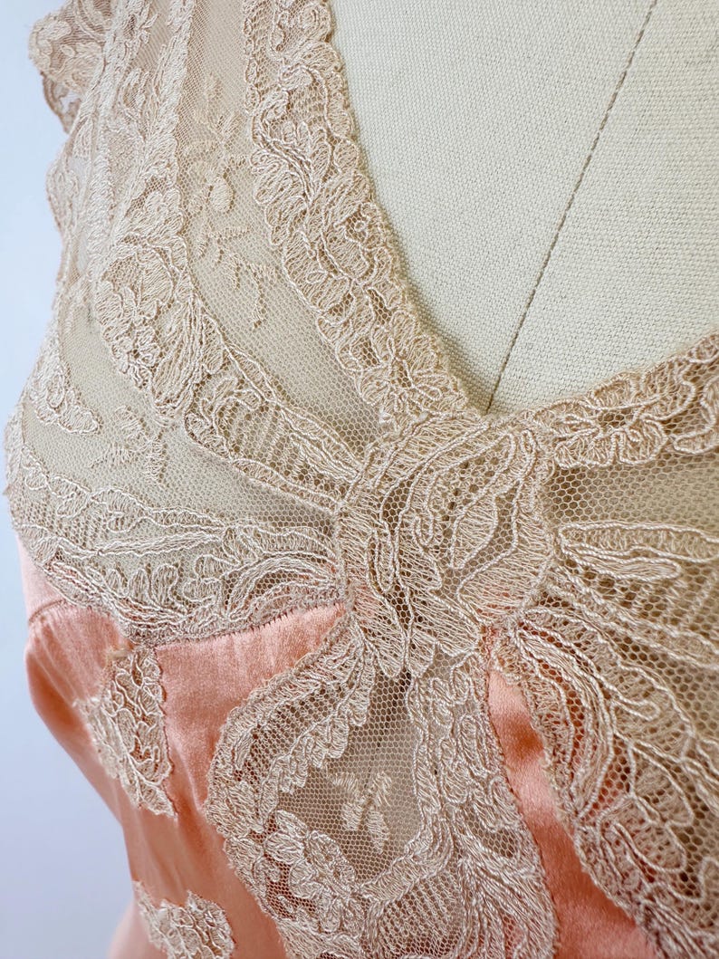 May include: Close-up of a peach-colored satin garment with intricate, cream-colored lace detailing. The lace overlay features floral and geometric patterns, creating a delicate and elegant texture. The garment appears to be a top or dress, showcasing a vintage or romantic aesthetic.