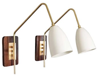 May include: A pair of wall sconces with a mid-century modern aesthetic. They have dark wood bases, brass details, and white, cone-shaped shades. The arms extend from the base, supporting the shades. A stylish lighting fixture.