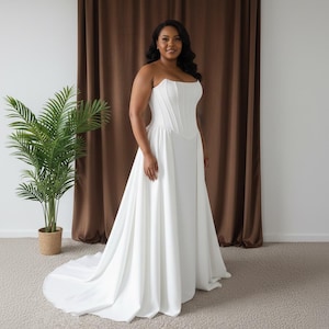 Puede incluir: Un vestido de novia blanco sin tirantes con un corpiño de corsé y una falda fluida. El vestido tiene una larga cola. Una persona lleva el vestido. Una planta en maceta está a la izquierda y una cortina marrón al fondo.