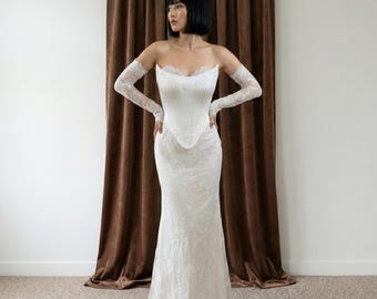 May include: A white, strapless wedding gown with a fitted bodice and a flowing skirt. The dress is adorned with delicate lace detailing. Matching lace gloves extend to the elbows. The gown is displayed in front of a brown curtain.