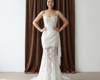 May include: A strapless white lace wedding dress with a corset bodice and a flowing skirt. The dress features intricate floral lace detailing and a draped fabric accent at the waist. The dress is floor-length.