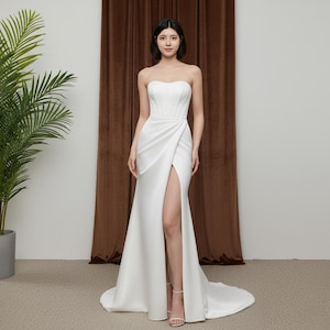 Puede incluir: Un vestido de novia blanco sin tirantes con un corpiño ajustado y una abertura alta. El vestido tiene una falda fluida que se arrastra por detrás. La modelo lleva tacones blancos con tiras. El fondo es una cortina marrón y una planta en maceta.