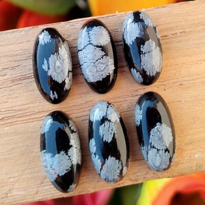 May include: Six oval-shaped snowflake obsidian cabochons. Each polished stone is black with white and gray snowflake-like patterns. The stones are arranged on a wooden surface, showcasing their glossy finish and unique natural designs.