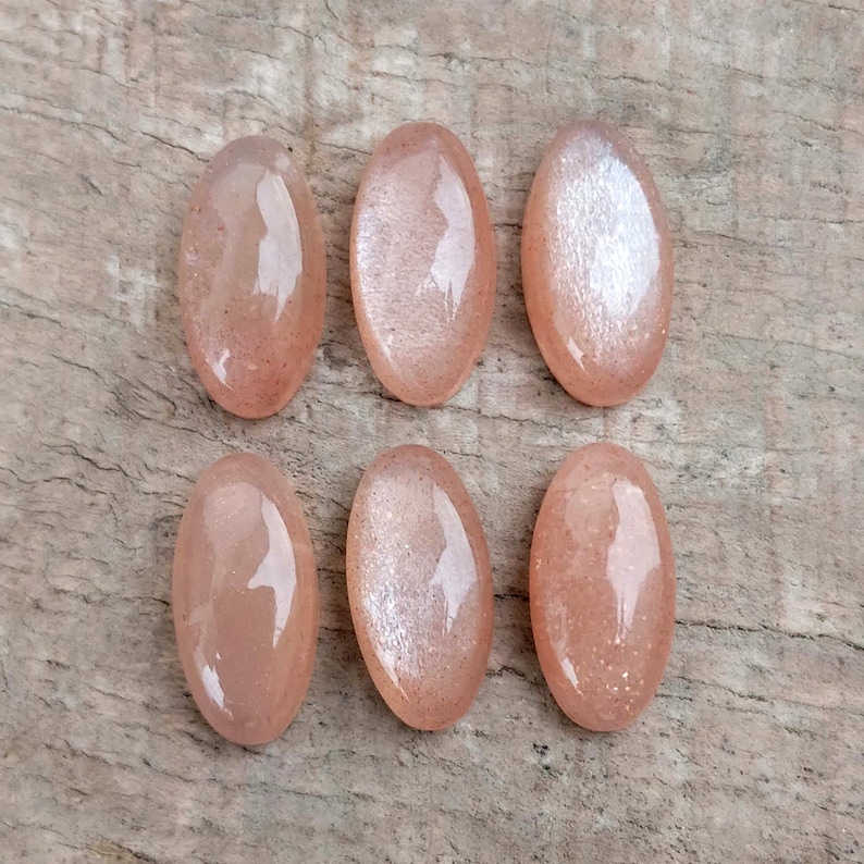 May include: Six oval-shaped, polished gemstones in varying shades of peach and pink are arranged on a wooden surface. The stones have a glossy finish, reflecting light, and are likely sunstone or similar. The gemstones are for jewelry making.