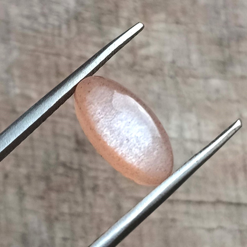 May include: A polished, oval-shaped gemstone, held by tweezers. The gem has a peach color with a subtle shimmer. The tweezers are silver-colored and the background is a light brown wooden surface.