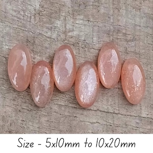 May include: Six oval-shaped, peach-colored gemstones with a shimmering, speckled appearance are arranged on a wooden surface. The text "Size - 5x10mm to 10x20mm" is at the bottom of the image. These stones could be used for jewelry making.