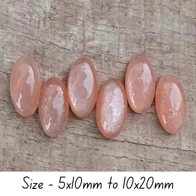 May include: Six oval-shaped, peach-colored gemstones with a shimmering, speckled appearance are arranged on a wooden surface. The text "Size - 5x10mm to 10x20mm" is at the bottom of the image. These stones could be used for jewelry making.