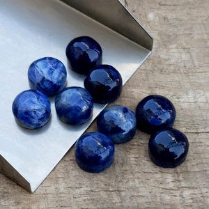 May include: A collection of nine polished, round, deep blue gemstones with varying patterns and textures. The stones are displayed on a silver metal surface and a wooden background. The gemstones have a glossy finish and reflect light.