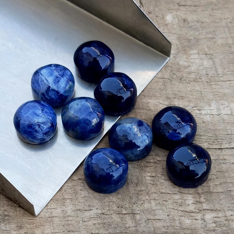 May include: A collection of nine polished, round, deep blue gemstones with varying patterns and textures. The stones are displayed on a silver metal surface and a wooden background. The gemstones have a glossy finish and reflect light.