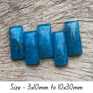 May include: Five rectangular, translucent, teal-coloured gemstones are arranged on a weathered wooden surface. The stones vary in size, with dimensions ranging from 3x10mm to 10x30mm. The image includes the text "Size - 3x10mm to 10x30mm".