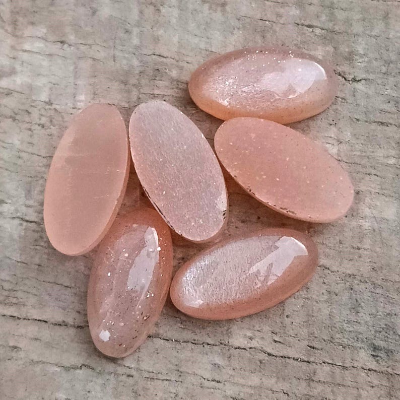 May include: Six oval-shaped, peach-colored gemstones with a polished, translucent appearance. The stones have a subtle sparkle and are arranged on a weathered, gray wooden surface. These gemstones are ideal for jewelry making.