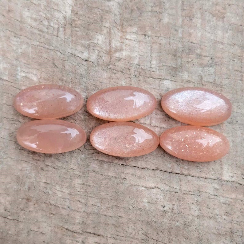 May include: Six oval-shaped, peach-colored gemstones with a shimmering, glittery appearance. The stones are arranged in two rows of three, each with a smooth, polished surface. The gemstones are likely used for jewelry making.