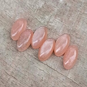 May include: Six oval-shaped, light peach-colored resin cabochons with glitter accents. The cabochons are arranged on a weathered wooden surface. The resin pieces have a glossy finish and are slightly translucent.