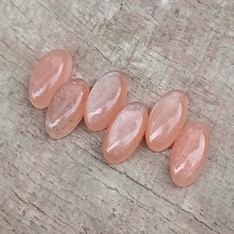 May include: Six oval-shaped, light peach-colored resin cabochons with glitter accents. The cabochons are arranged on a weathered wooden surface. The resin pieces have a glossy finish and are slightly translucent.