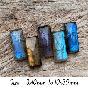 May include: Five rectangular labradorite gemstones in shades of blue, purple, and gold are displayed on a wooden surface. The stones vary in size, with dimensions ranging from 3x10mm to 10x30mm. The text "Size - 3x10mm to 10x30mm" is at the bottom.