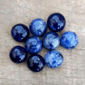 May include: Nine polished, round sodalite gemstones in varying shades of blue, from deep navy to lighter, marbled hues. The stones are arranged on a weathered, light brown wooden surface, showcasing their glossy finish and natural patterns.
