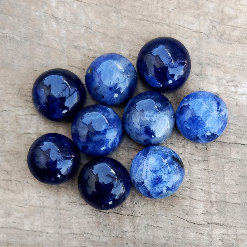May include: Nine polished, round sodalite gemstones in varying shades of blue, from deep navy to lighter, marbled hues. The stones are arranged on a weathered, light brown wooden surface, showcasing their glossy finish and natural patterns.
