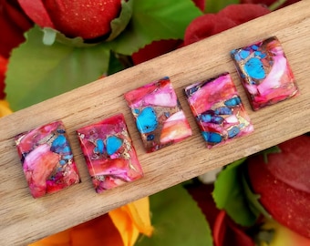 May include: Five rectangular gemstone cabochons in shades of pink, red, blue, and gold. The stones are displayed on a wooden surface, with a floral backdrop of red and green.