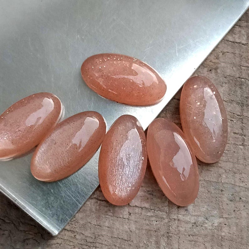 May include: Six oval-shaped, peach-colored gemstones with a shimmering, speckled appearance. The polished stones are arranged on a silver surface, with a wooden background visible. These gemstones are likely for jewelry making.