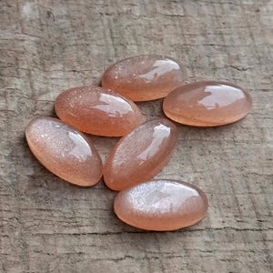 May include: Six oval-shaped, peach-colored gemstones with a shimmering, speckled appearance. The polished stones are arranged on a weathered, wooden surface, showcasing their translucent quality and subtle variations in color and texture. These gemstones are ideal for jewelry making.