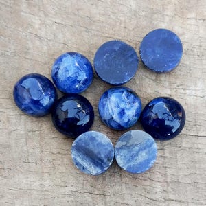 May include: A collection of blue and white polished stones and flat, round, dark blue stones. The stones are arranged on a weathered wooden surface. The polished stones have a glossy finish, while the flat stones have a matte appearance.