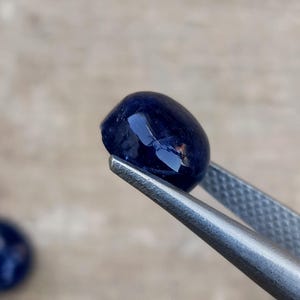 May include: A polished, oval-shaped, deep blue gemstone is held by a pair of silver tweezers. The gem has a smooth, reflective surface, catching the light. The background is blurred, emphasizing the gem's color and shape.