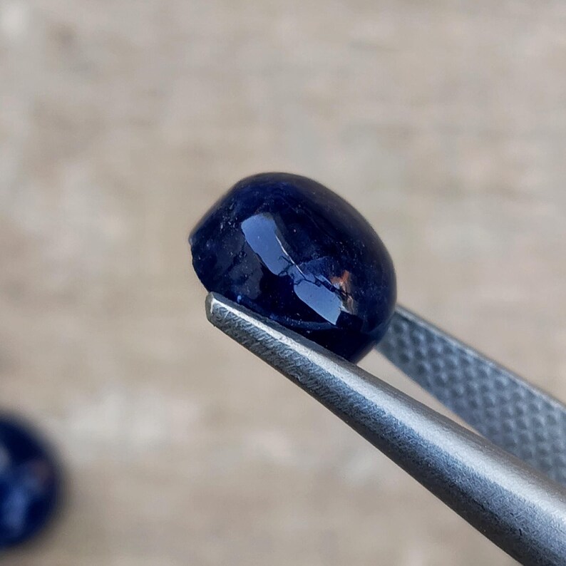 May include: A polished, oval-shaped, deep blue gemstone is held by a pair of silver tweezers. The gem has a smooth, reflective surface, catching the light. The background is blurred, emphasizing the gem's color and shape.
