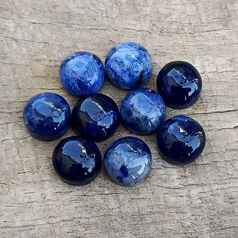 May include: Nine polished, round gemstones in shades of blue, from deep navy to lighter, marbled hues. The stones are arranged on a weathered, wooden surface, showcasing their glossy, reflective surfaces.