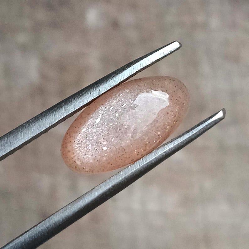 May include: A polished, oval-shaped gemstone held by tweezers. The stone has a peach color with sparkling inclusions. The gem is approximately 0.75 inches in length, and is likely a sunstone or similar variety.