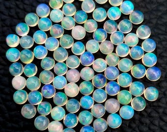 May include: A collection of round, iridescent opal gemstones. The opals display a range of colors, including blue, green, and white, with a shimmering effect. The gemstones are arranged closely together on a dark surface.