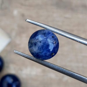 May include: A round, polished sodalite gemstone held by tweezers. The gemstone is a deep blue color with white and gray marbling. The surface is smooth and reflective, with small dark inclusions. The background is a blurred, neutral tone.