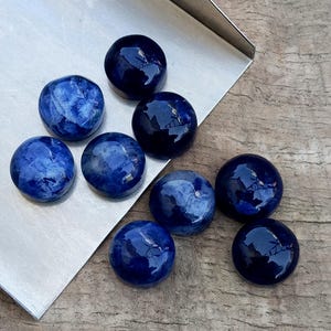 May include: A collection of eight polished, round, blue gemstones with varying patterns and shades. Some stones have a marbled appearance, while others are a solid, deep blue. The gemstones are displayed on a silver tray and a wooden surface.