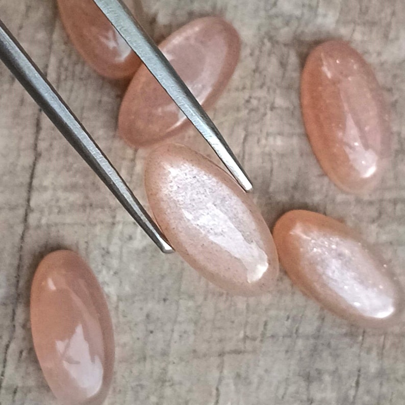 May include: Close-up of several oval-shaped, peach-colored gemstones. One gemstone is held by silver tweezers. The gemstones have a subtle shimmer and are arranged on a light brown surface. The gemstones are likely for jewelry making.