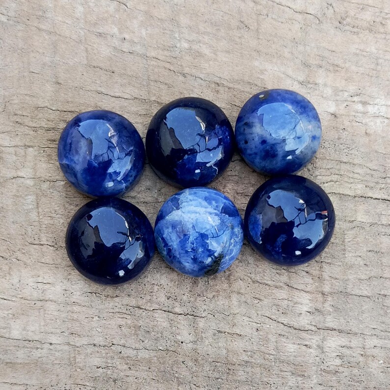 May include: Six polished, round, blue sodalite gemstones. The stones vary in shades of deep blue with white marbling. The gemstones are arranged on a weathered, light brown wooden surface.