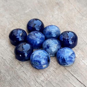 May include: A collection of nine polished, round sodalite gemstones. The stones vary in shades of blue, from deep navy to lighter, marbled hues. They are arranged on a weathered, light brown wooden surface.