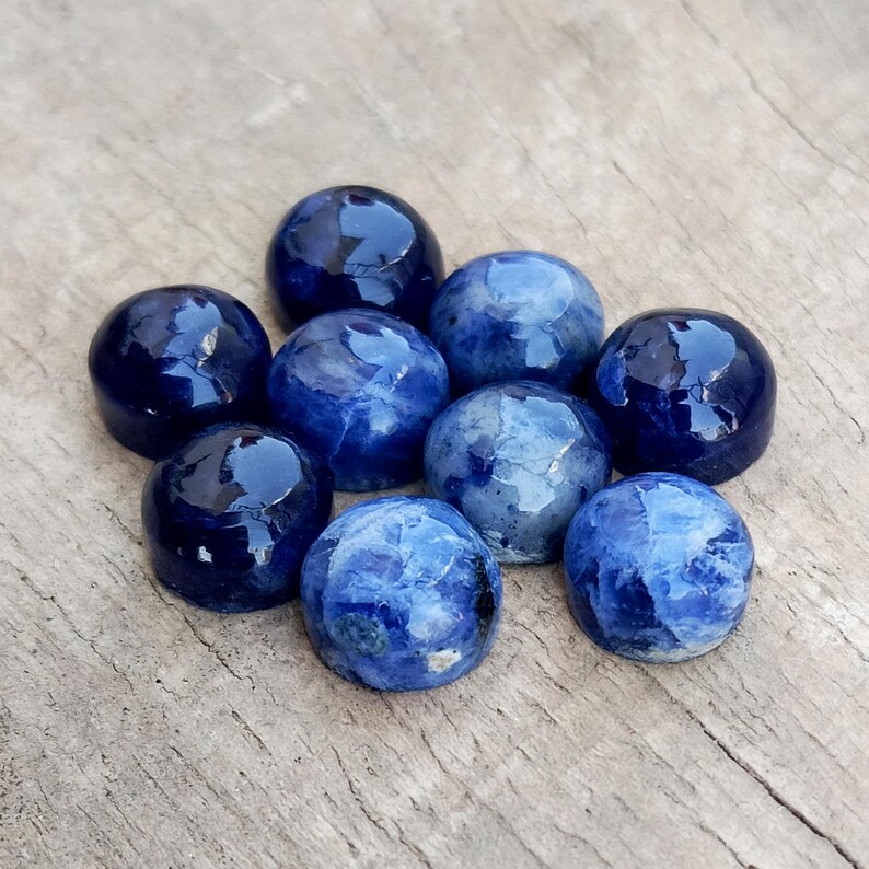 May include: A collection of nine polished, round sodalite gemstones. The stones vary in shades of blue, from deep navy to lighter, marbled hues. They are arranged on a weathered, light brown wooden surface.