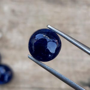 May include: A deep blue, polished gemstone held by silver tweezers. The gem is round with a smooth, reflective surface. The background is a blurred, neutral tone, highlighting the gem's color and shape.